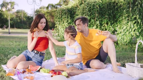 Family Enjoys a Sunny Picnic in a Park While Sharing Fruit and Laughter on a Warm Afternoon