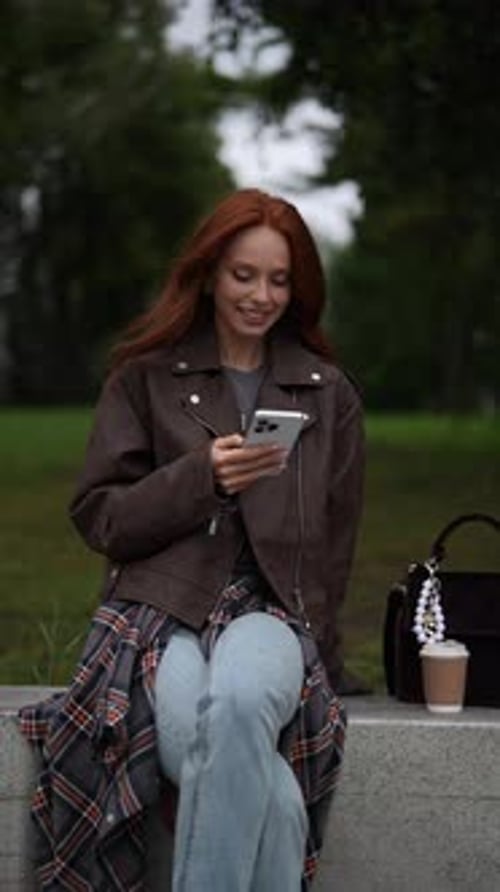 Vertical Portrait of Cheerful Redhead Female Smiling While Texting Using Phone Sitting on Park Bench