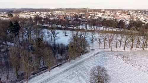 Snowy rural landscape with open fields scattered leafless trees and distant town featuring church