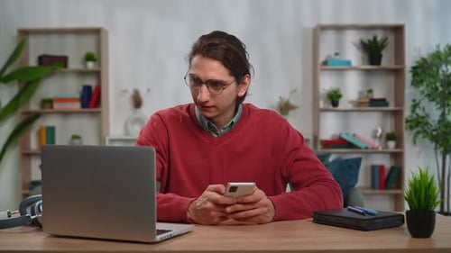 Young Adult Man Spending Time at Home Man Student Sits at Desk Works on Laptop Talking Online By