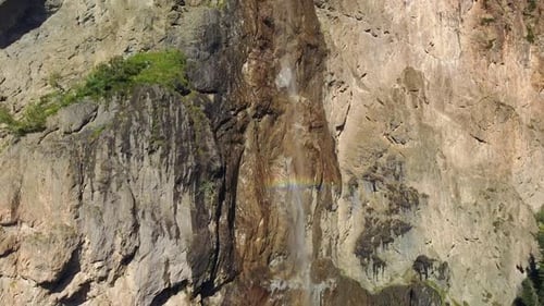 alpine waterfall from the rocky wall with rainbow colors, aerial bottom to top shot