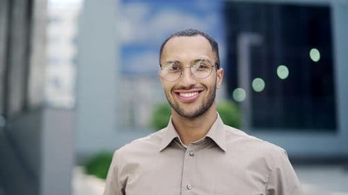Close up portrait of a young adult man in a shirt and glasses smiling and looking at the camera.