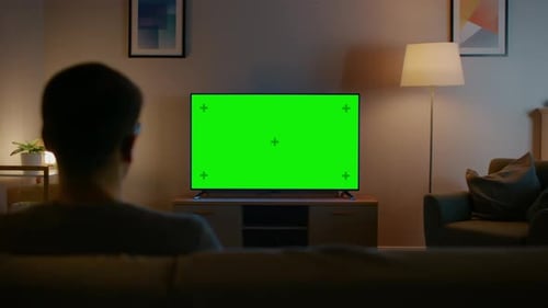 Young Man in Glasses is Sitting on a Sofa and Watching TV with Horizontal Green Screen Mock Up. It'