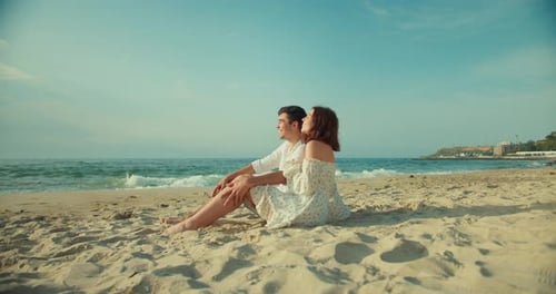 Happy Couple a Brunette Guy and a Girl in a White Dress are Sitting on the Beach Talking During