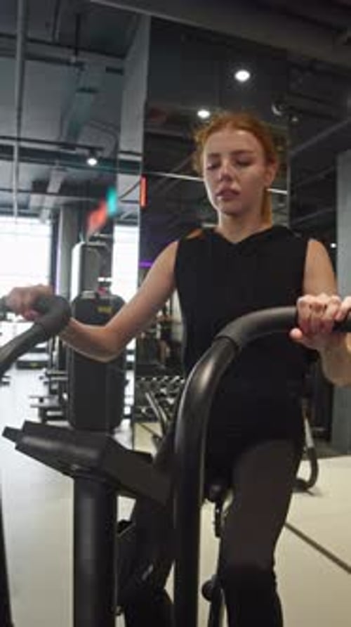 A Young Woman is Exercising Her Legs on an Exercise Bike at the Gym Holding on to the Handles for