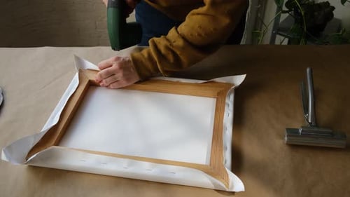 Artist's Hands Stretching Canvas on Stretcher Bars Using Staple Gun in Workshop
