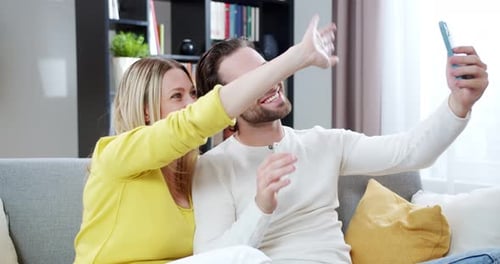 Caucasian Happy Couple Laughing and Sitting on Couch While Using Smartphone and Videochattin Inside