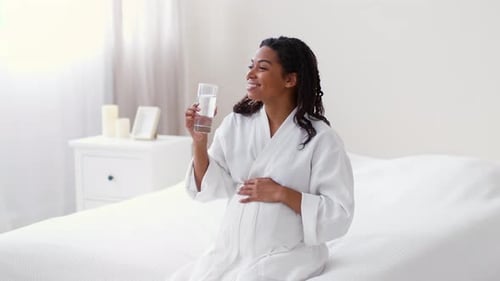 Schwangere Frau genießt ein erfrischendes Getränk, während sie auf einem Bett in einem hellen Raum sitzt