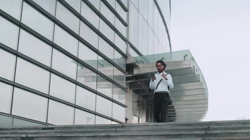 Young Asian Businessman in a White Shirt Walks Past the Business Center with a Smartphone and a