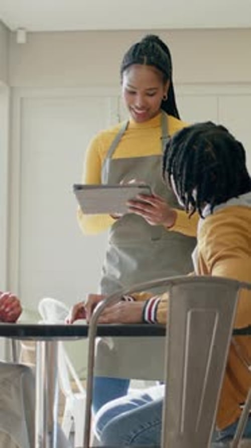 Vertical video: Approaching server in gray apron tapping tablet, showing menu at cafe, helping man