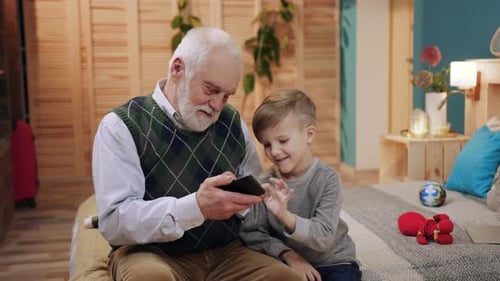 Grandfather and Little Boy Sitting on Bed and Looking Directly at Screen of Smartphone in Background