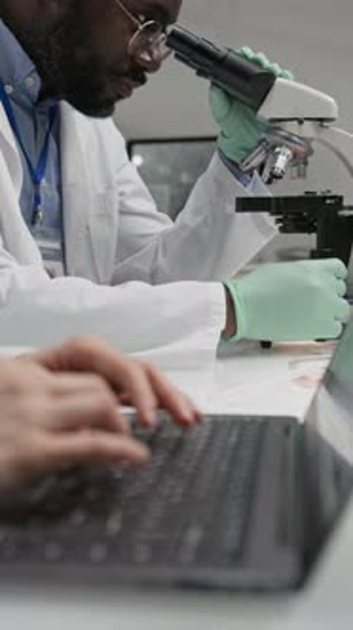 Scientist Uses Microscope While Another Types on Laptop