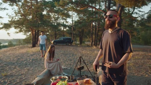 Stylish Hipster in Glasses is Relaxing with Friends on the River Bank and is Cooking on a Fire While