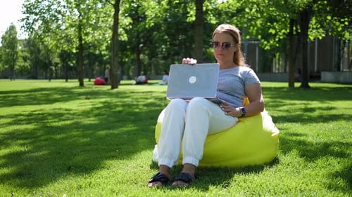 Une jeune femme avec un ordinateur portable est assise dans un fauteuil dans un parc moderne