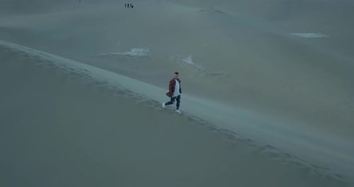 Aerial follow shot of a boy walking alone in a barren desert on a large sand dune during a windy