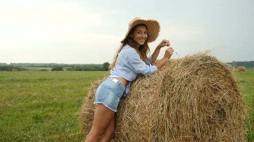 Pretty Young Woman Stands Near Haystack And Plays With Straw. Farmer's Wife Walks On The Field