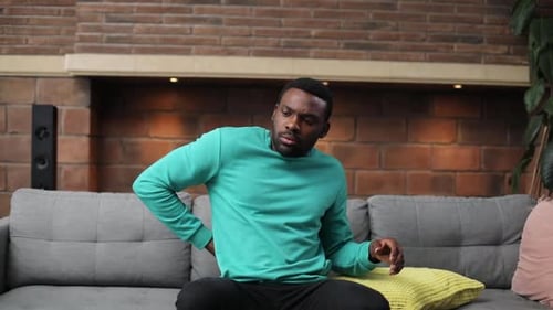 Likable African Guy Sitting on Sofa with Colored Pillows in Living Room Looking at Screen of