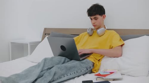 Young Person Engages with Laptop While Seated on Bed in Cozy Room Wearing Headphones and Focused on