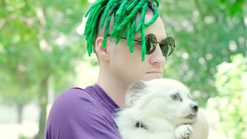Young Man with Green Dreadlocks in Sunglasses Holding Fluffy White Dog in Hands Stroking Kissing Pet