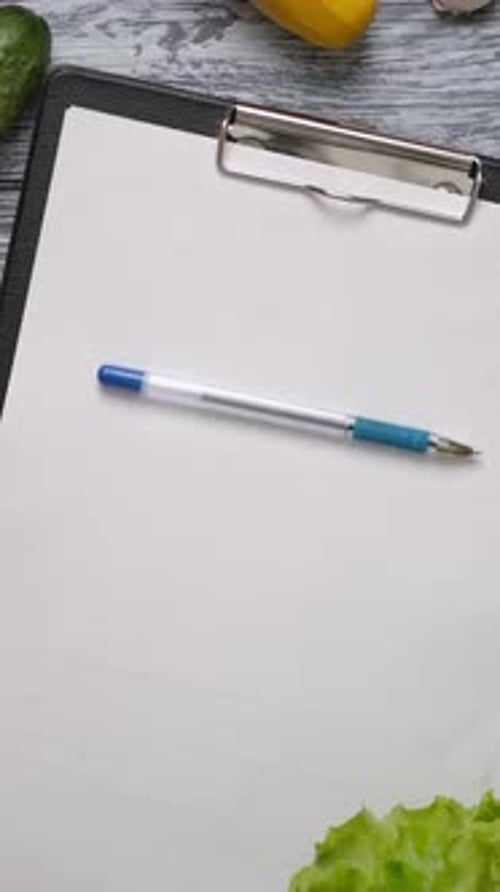 Vertical of Pen Rolling on Blank Clipboard on Kitchen Table with Vegetables