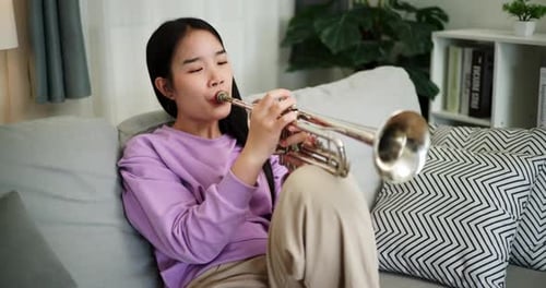 A young woman practices playing the trumpet while sitting on a sofa in a cozy living room.
