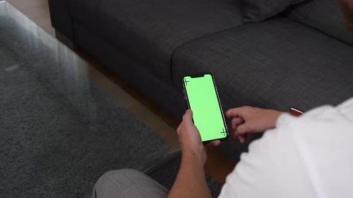 Male model swipes over screen of his smartphone while sitting on sofa in living room. Green screen p