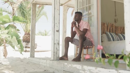 Thoughtful african american man sitting on porch of wooden beach house looking away, slow motion