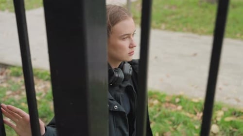 Pensive Young Woman Standing Behind Metal Park Fence