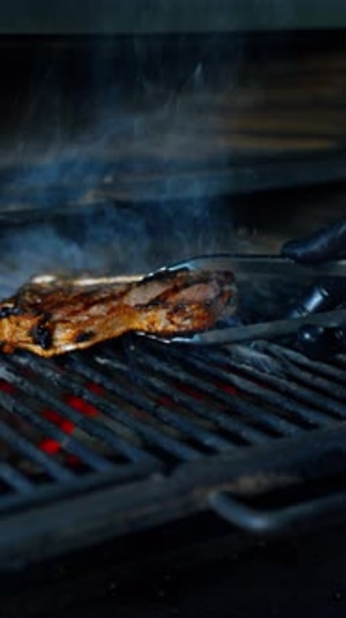 Meat on the bone is cooked on the grill. Gloved hand turns the piece of beef with tongs.