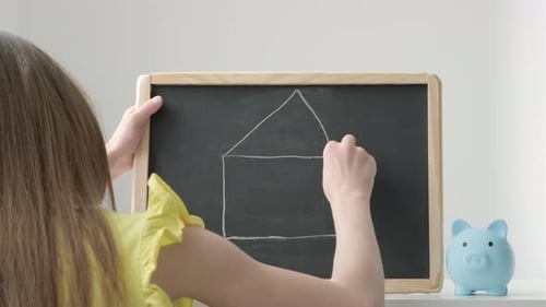 Child Drawing House on Chalkboard Next to Piggy Bank