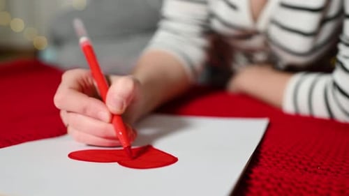 Young Woman in a Striped Sweater Draws a Red Heart on a White Valentine Card Sheet and Lies on a Red