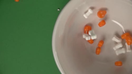 Zenith view of orange skulls and bones shaped candy falling on a plate, on a chroma background.
