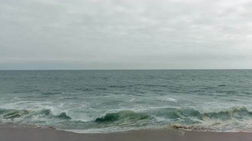 Niedrige Luftaufnahme vom Sandstrand, der sich in Richtung schaumiger, brechender Meereswellen und düsterer Himmel mit Meer bewegt
