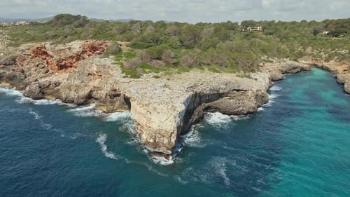 A rocky cliff juts into the turquoise sea, surrounded by lush forest and rugged coastline. The aeria