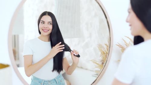Smiling young woman looking at mirror, combing long dark hair and getting ready for a great day.