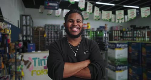 Man in a black apron smiling with arms crossed in a paint store, surrounded by shelves filled