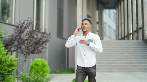 cheerful african american guy walks and talks urban city street in casual clothes using mobile phone