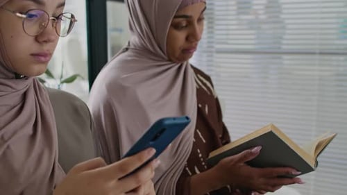 Two Girls in Hijabs Using Phone and Reading Book Before Appointment in Clinic