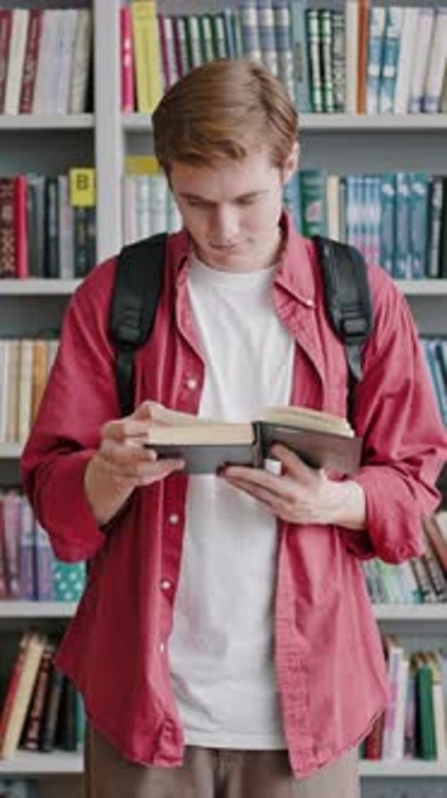 Young Man Reads Book Standing Against Bookshelves in Library