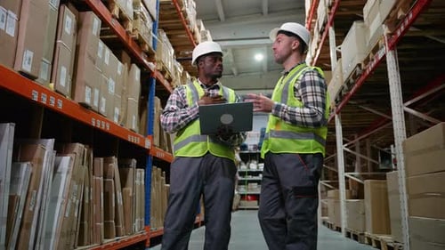 Two Warehouse Employees Wearing Safety Vests and Helmets Discuss Logistics While Working on a Laptop