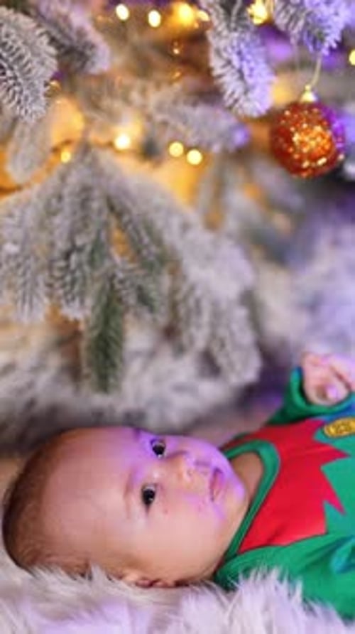 Beautiful infant baby wearing elf green costume lies under Christmas tree.