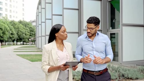 Two diverse business colleagues walking outside an office building and reviewing a tablet