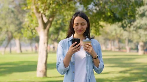 Woman Uses Phone and Drinks Coffee in Park