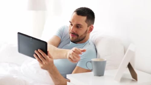 Man in bed uses tablet, drinks coffee