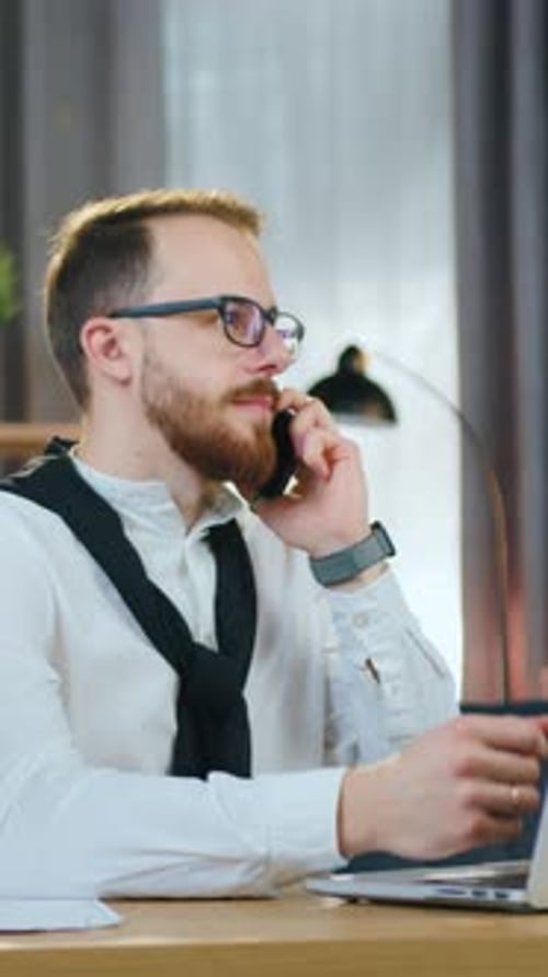 Sympathischer Mann, Mann mit Brille, sitzt an seinem Arbeitsplatz im modernen Wohnzimmer und spricht auf dem Smartphone