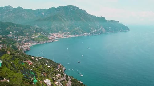 Aerial view of a coastal town with a prominent cliffside park, surrounded by blue sea and mountains,