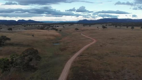 Aerial view of a dirt road that extends through a field of grass and towards some high mountains tha
