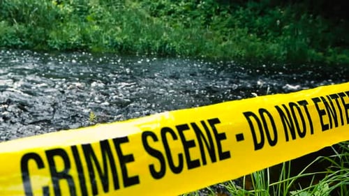Yellow Crime Scene police tape pulled infront of a fast flowing rural river shore during cloudy day.