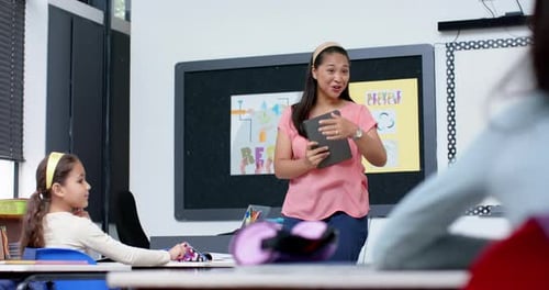 Teacher in classroom holding tablet, smiling at students during school lesson