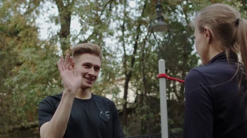 Young Smiling Caucasian Sportsman Takes High Five to Young Caucasian Sportswoman on a Workout Area